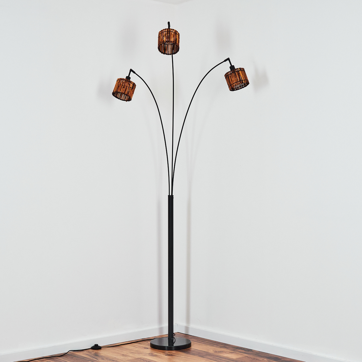 Eine Stehlampe mit schwarzem Ständer und drei gebogenen Armen, die jeweils einen gewebten Lampenschirm tragen, steht in einer Zimmerecke.