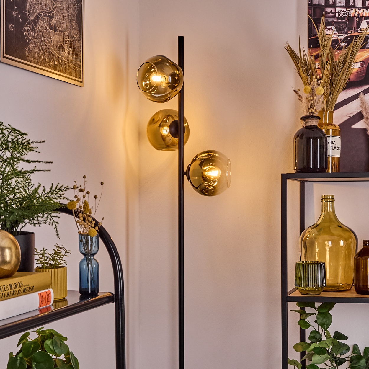 Moderne Lampe mit drei goldenen Glaskugeln. Regale mit Vasen und Pflanzen sind sichtbar. Eine Karte und ein Poster sind an der Wand.
