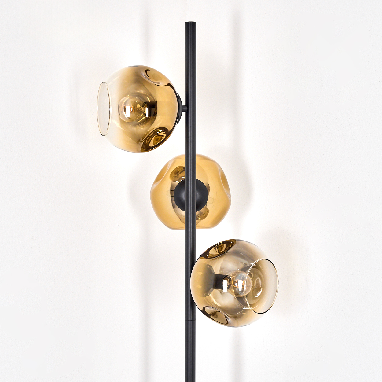 Eine moderne Stehlampe mit drei bernsteinfarbenen Glaskugeln. Die Lampe hat ein schwarzes Metallgestell vor einer weißen Wand.