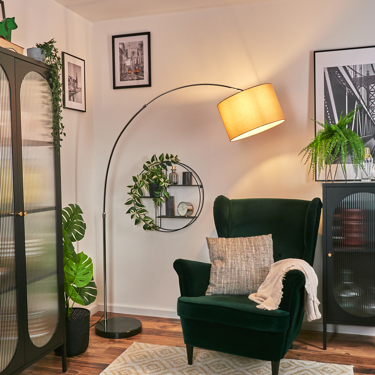 Ein dunkelgrüner Sessel mit Kissen und Decke. Eine schwarze Stehlampe mit beigem Lampenschirm. Schwarze Schränke und Bilder an der Wand.