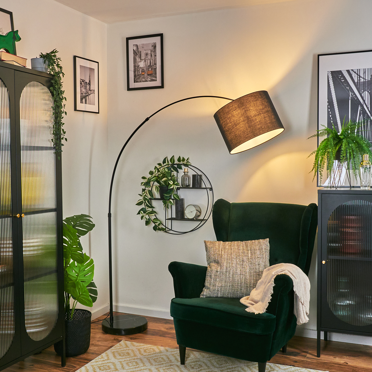 Ein grüner Sessel mit Kissen und Decke steht neben einer schwarzen Stehlampe. Ein dekoratives Regal mit Pflanzen hängt an der Wand.