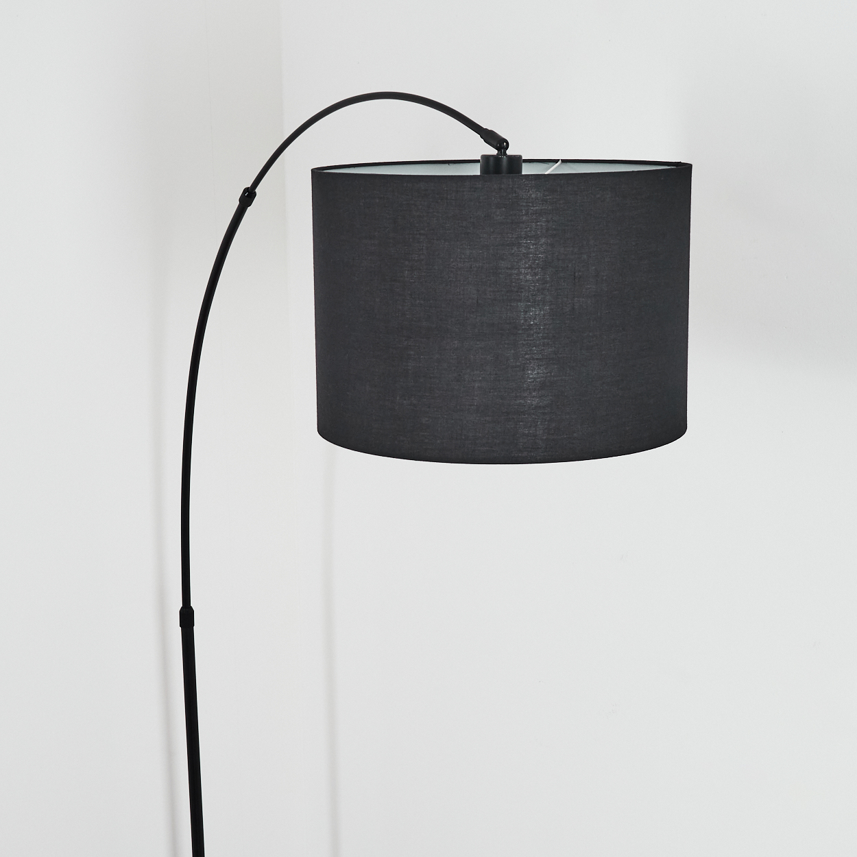 Eine schwarze Stehlampe mit gebogenem Arm und zylindrischem Lampenschirm vor einer weißen Wand.