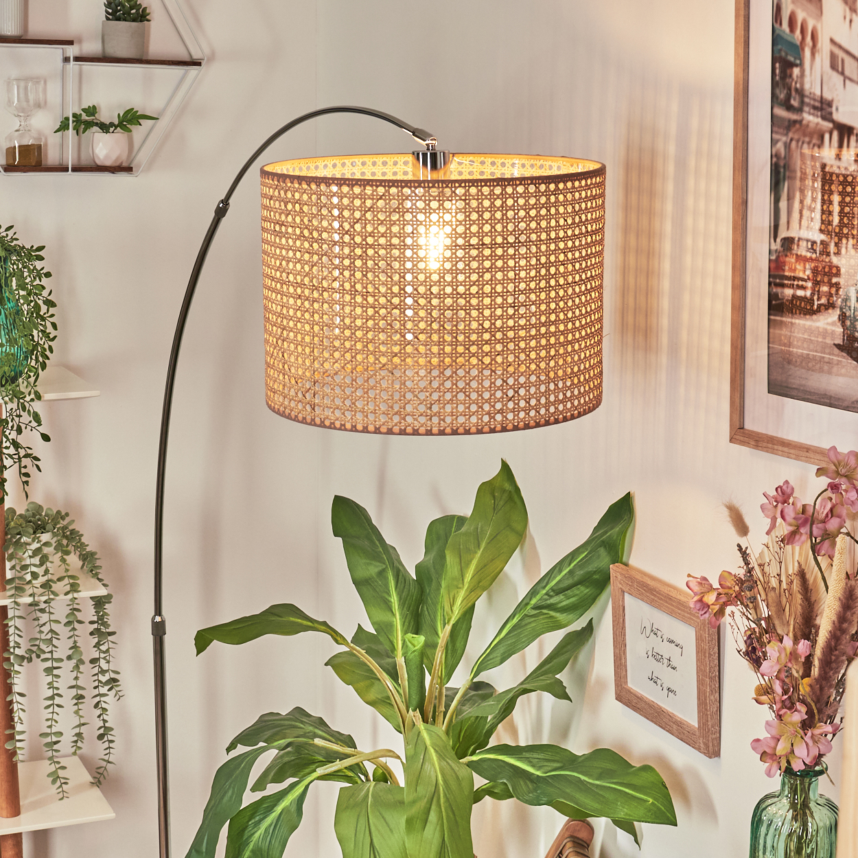 Eine Stehlampe mit gewebtem Schirm beleuchtet eine grüne Pflanze. Ein gerahmtes Bild und Blumen sind an der Wand zu sehen.