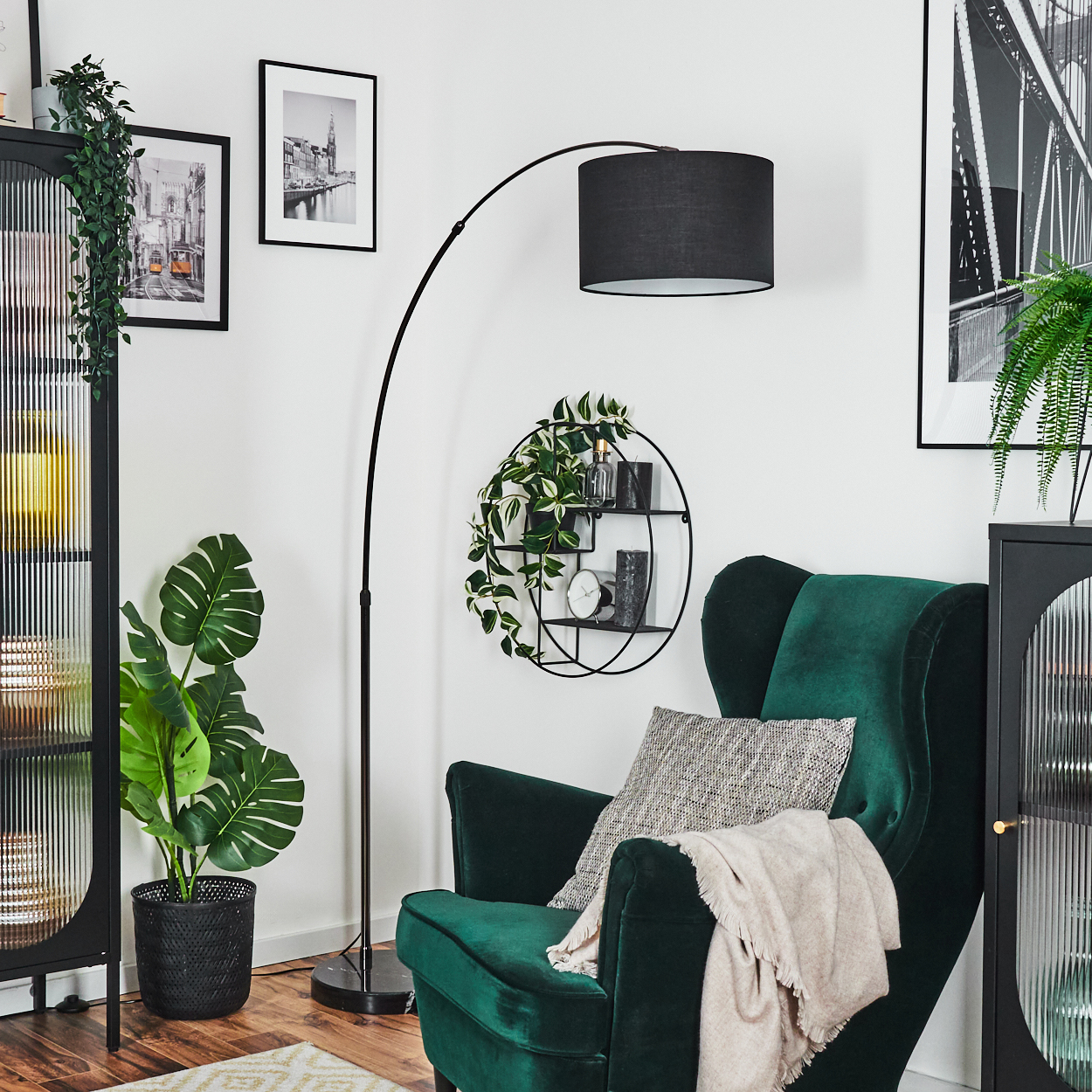 Ein grüner Samtsessel steht in einem Raum. Eine schwarze Lampe, Bilder, ein Regal mit Pflanzen und ein Schrank sind ebenfalls vorhanden.
