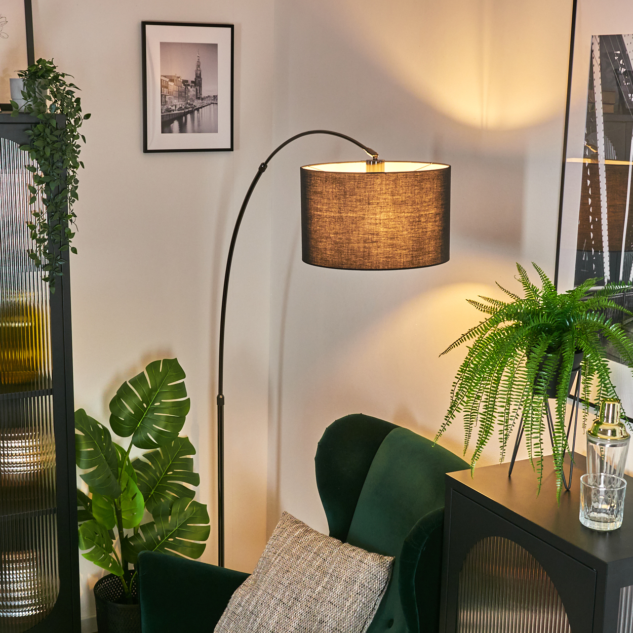 Eine Zimmerecke mit Stehlampe, grünem Stuhl, Pflanzen, gerahmter Kunst und einem schwarzen Schrank. Die Szene ist gut beleuchtet und stilvoll.