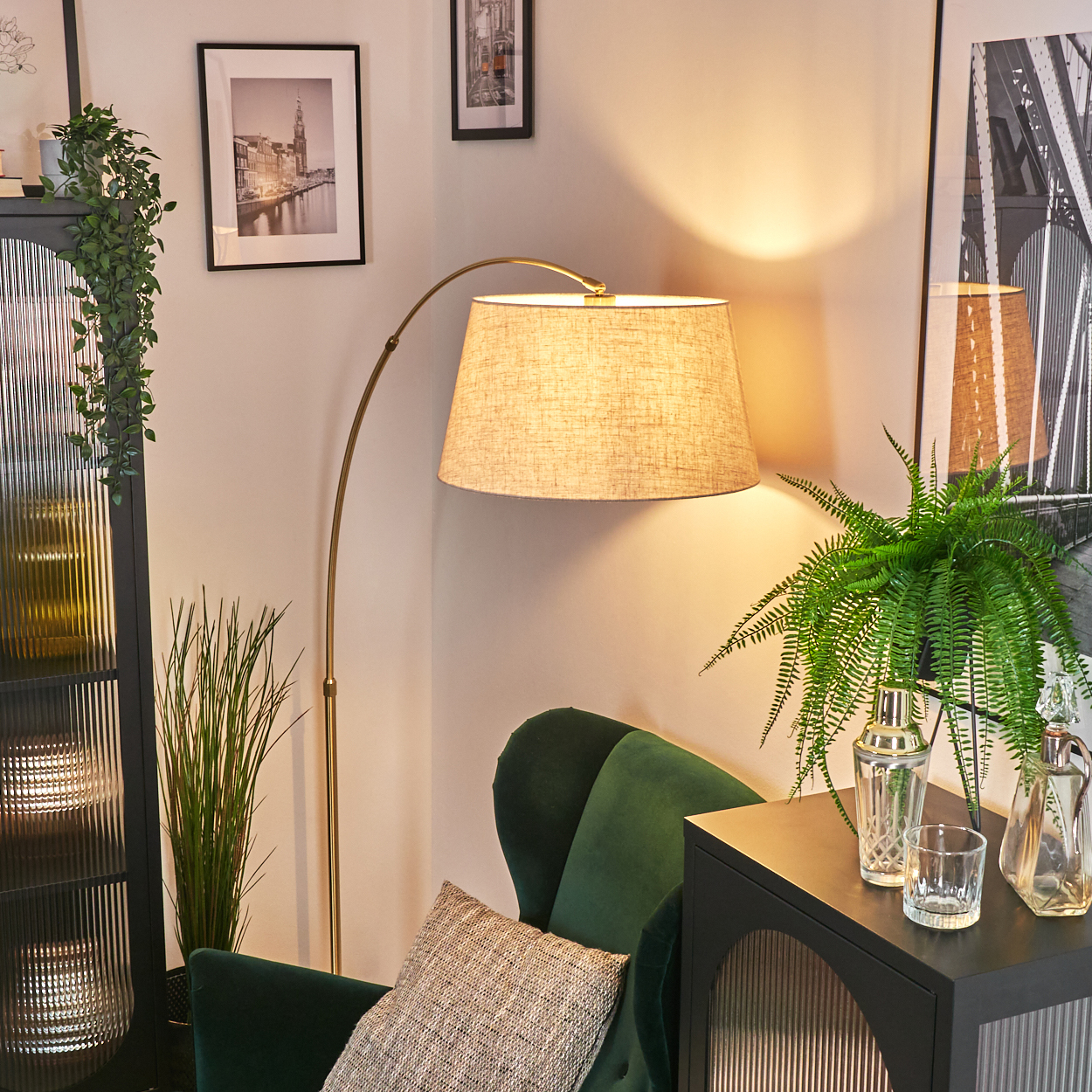 Ein Raum mit grünem Sessel, Lampe und Bildern an der Wand. Auf dem Tisch steht eine Pflanze, und auf dem Boden eine hohe Pflanze.