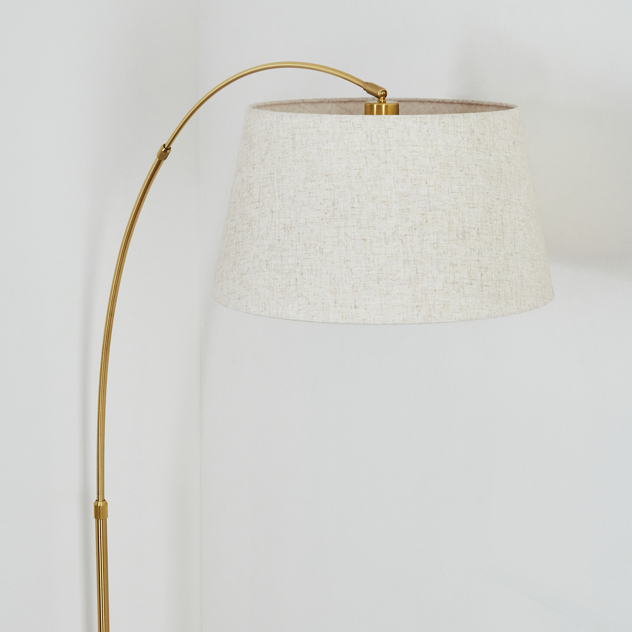 Eine goldene Stehlampe mit beigem Lampenschirm steht vor einer weißen Wand. Die Lampe hat einen gebogenen Arm.