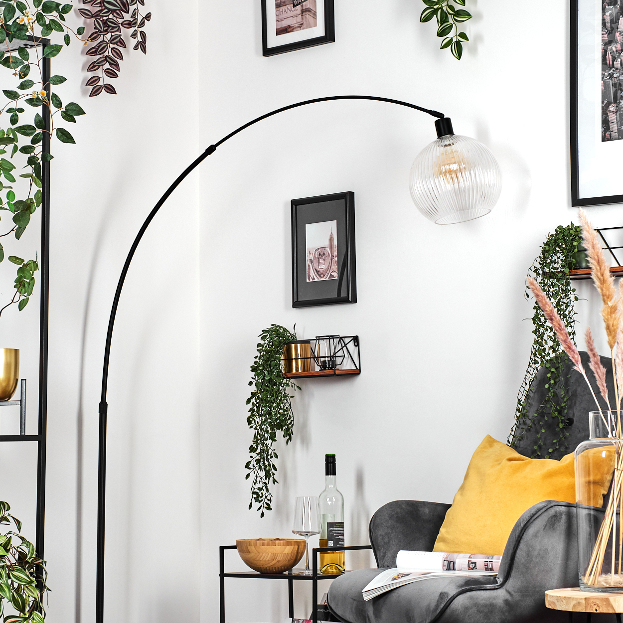 Ein modernes Wohnzimmer mit schwarzer Stehlampe, grauem Sessel, gelbem Kissen und dekorativen Pflanzen. Überkopf hängt eine Glaslampe.