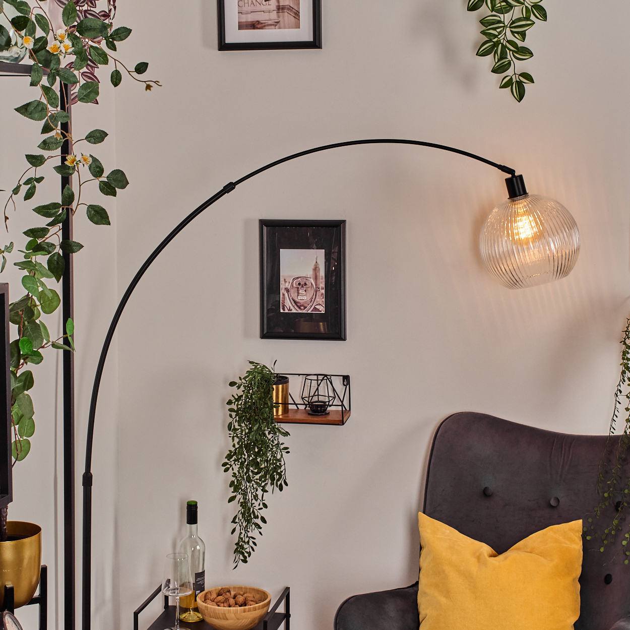 Ein modernes Wohnzimmer mit einer schwarzen Bogenlampe, gerahmten Bildern, einem grauen Sessel und Pflanzen.