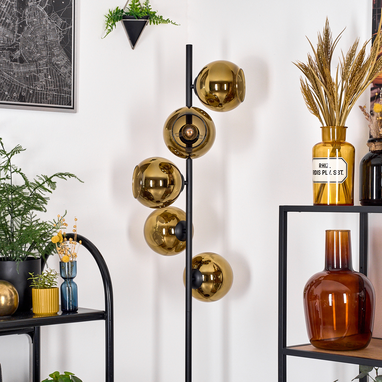 Eine moderne Lampe mit fünf goldenen Glaskugeln steht in einem Raum mit Regalen und Pflanzen. Eine Karte hängt an der Wand.