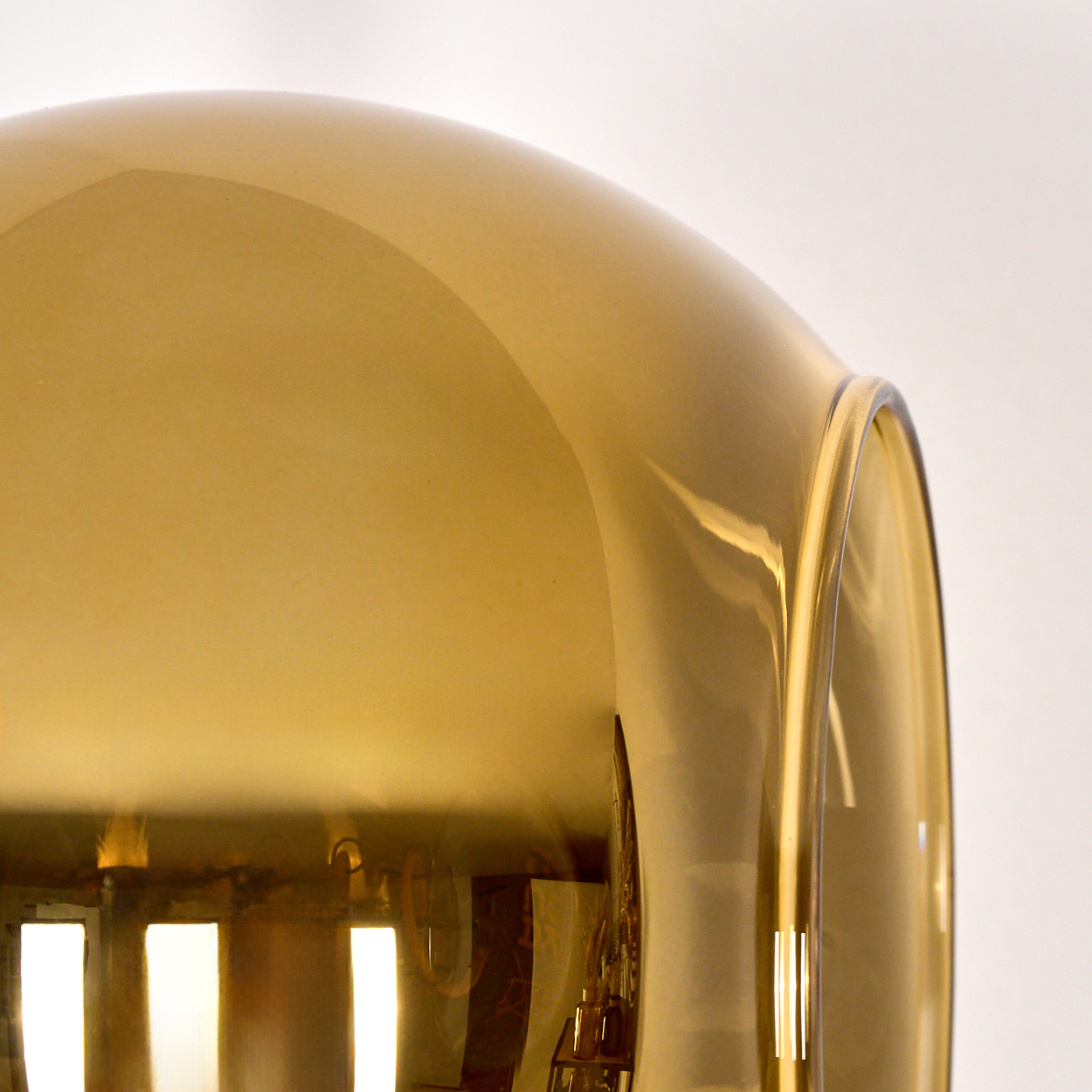 Nahaufnahme eines goldenen, gebogenen Glasobjekts vor weißem Hintergrund. Reflexionen und Licht spielen auf der Oberfläche.