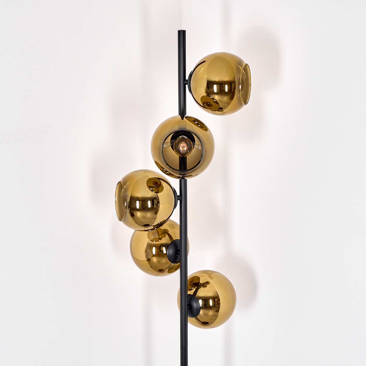 Eine moderne Stehlampe mit fünf goldenen Glaskugeln an einer schwarzen Metallstange vor einer weißen Wand.