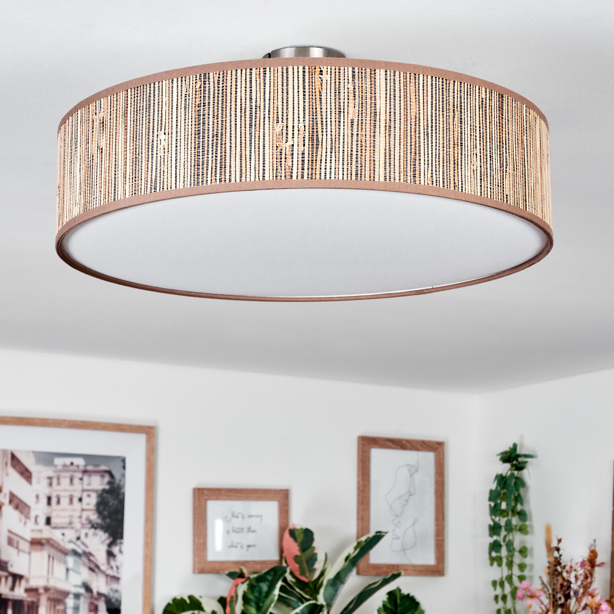 Deckenleuchte mit Rattan-Schirm und weißem Innenraum. Darunter gerahmte Kunst und Pflanzen.