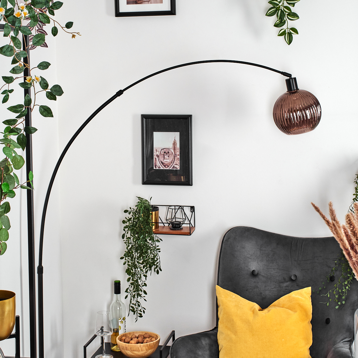 Wohnzimmerszene mit Lampe, Sessel mit gelbem Kissen, Zierpflanzen und gerahmten Bildern an einer weißen Wand.