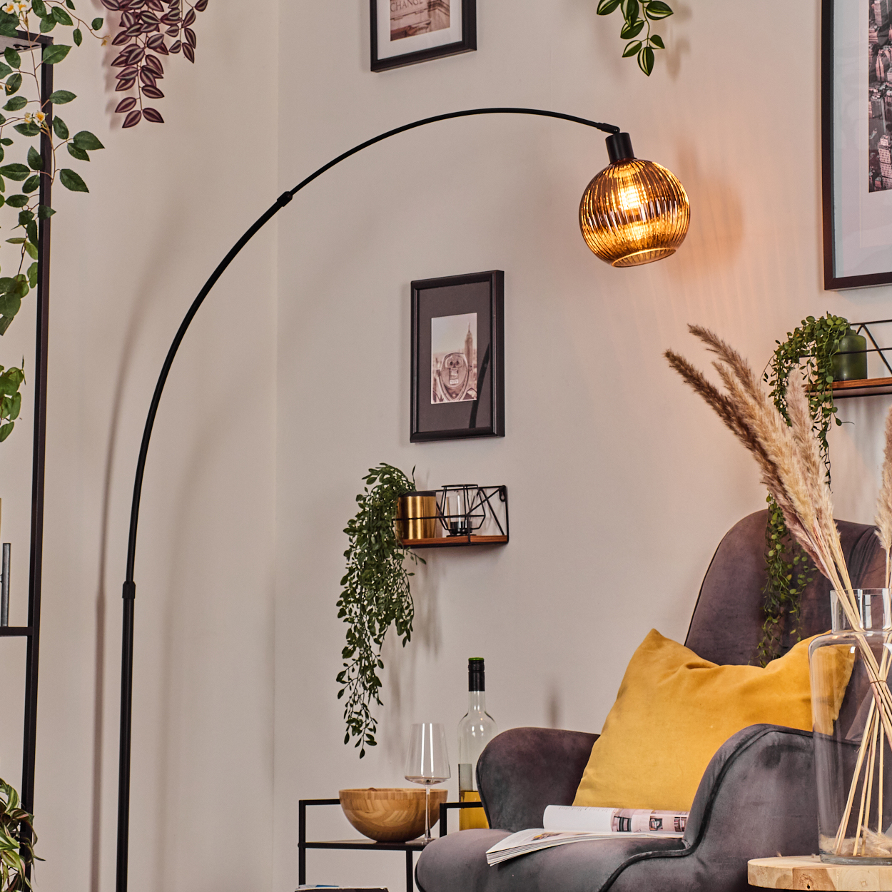 Ein modernes Wohnzimmer mit einer schwarzen Bogenlampe, einem grauen Sessel, einem gelben Kissen und Zierpflanzen. Eine Weinflasche ist sichtbar.