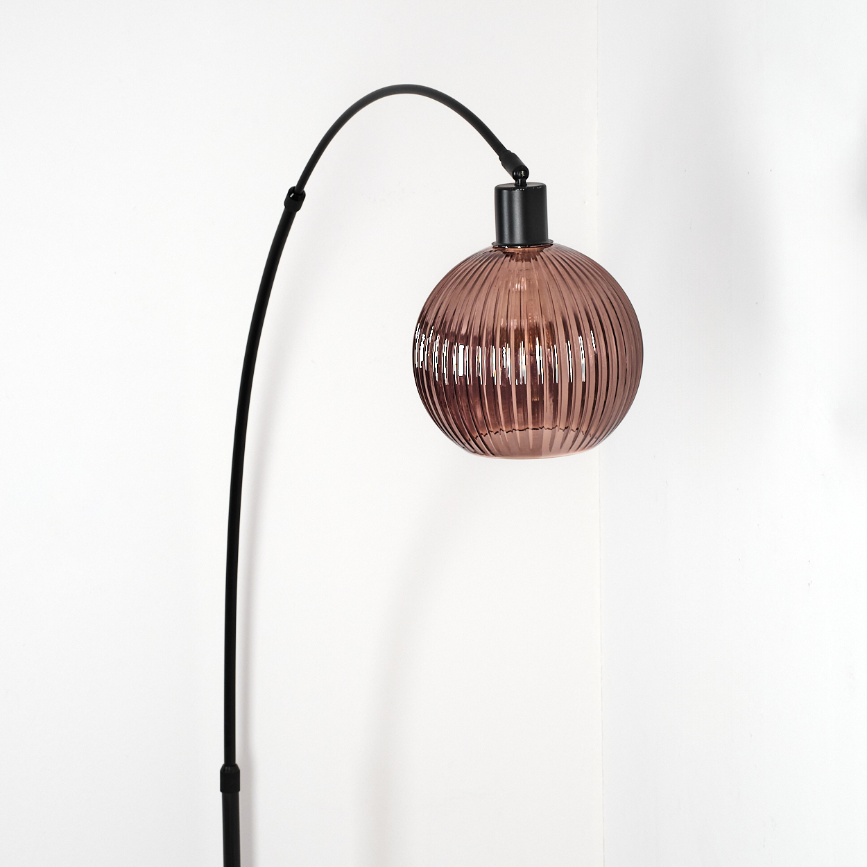 Eine moderne Lampe mit schwarzem, gebogenem Arm und rundem, geripptem, rosa-braunem Glasschirm, vor weißer Wand.