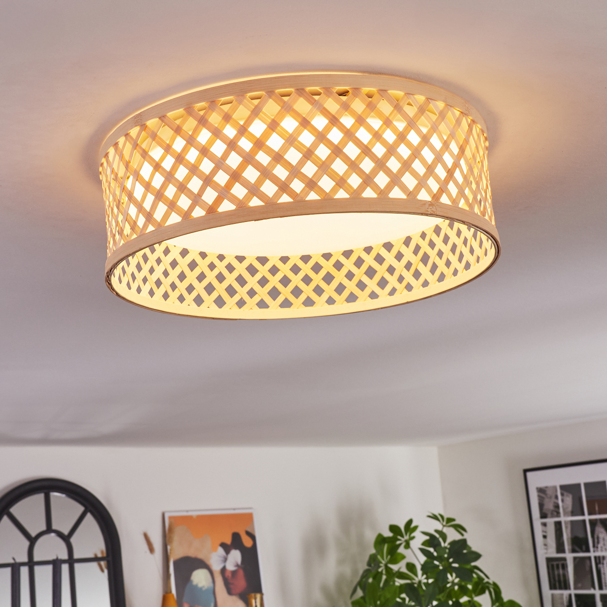 Eine Deckenlampe aus gewebtem Bambus ist beleuchtet. Die Lampe befindet sich über einem Raum mit Bildern und Pflanzen.