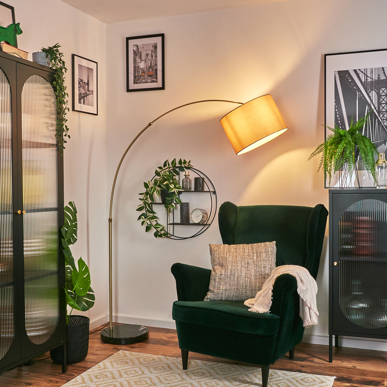 Ein grüner Sessel mit Kissen und Decke steht unter einer Lampe. Ein schwarzer Schrank und Bilder sind im Hintergrund.