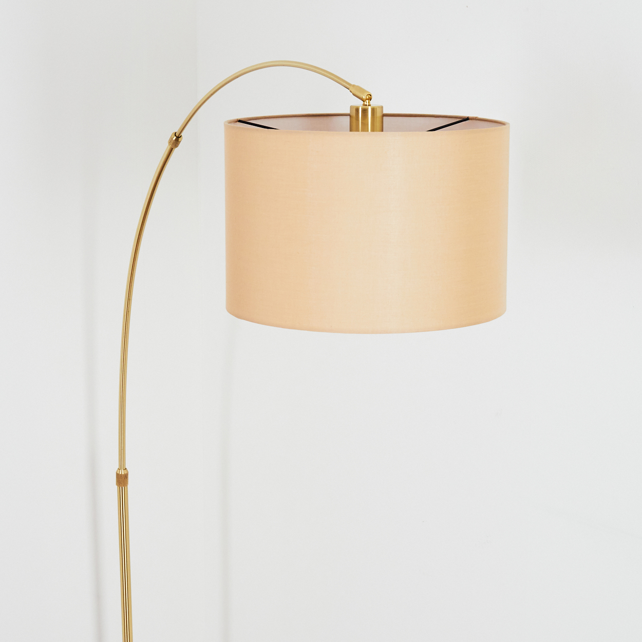 Eine goldene Stehlampe mit beigem Lampenschirm steht vor einer weißen Wand. Die Lampe hat einen gebogenen Arm.