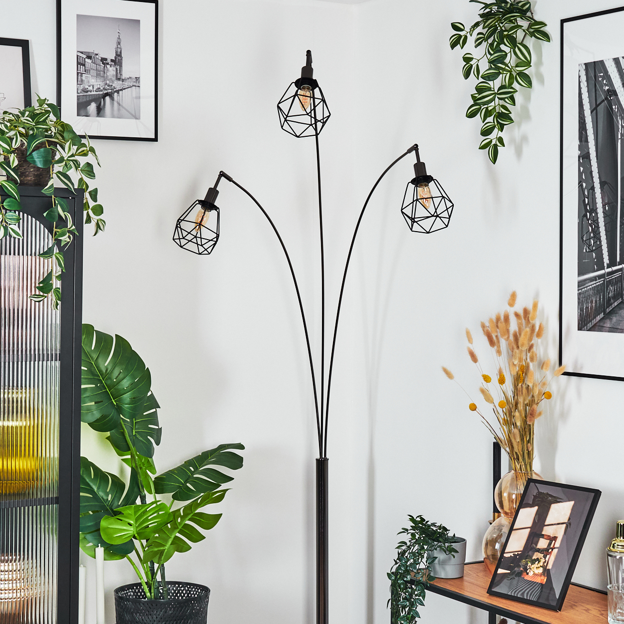 Ein modernes Zimmer mit einer schwarzen Stehlampe mit drei Lichtern, Pflanzen, Bildern und einem Holzregal mit Dekorationen.