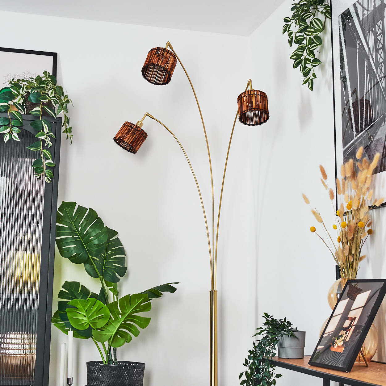 Ein modernes Wohnzimmer mit einer goldenen Stehlampe und drei Lampenschirmen. Grüne Pflanzen und ein gerahmtes Bild schmücken den Raum.