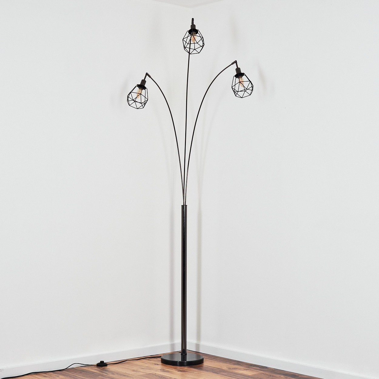 Eine schwarze Stehlampe mit drei Lichtern, jedes in einem geometrischen Käfig, steht in einer Ecke. Der Hintergrund sind weiße Wände und ein Holzboden.