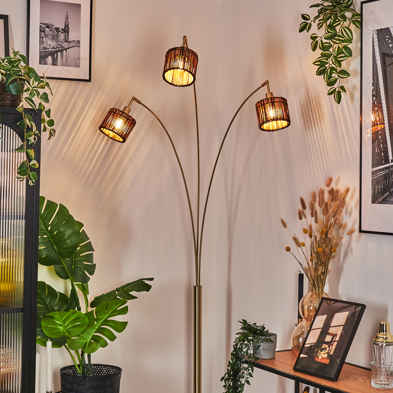 Eine Stehlampe mit drei Lampenschirmen beleuchtet einen Raum mit Pflanzen und gerahmten Bildern. Links und rechts sind Pflanzen.