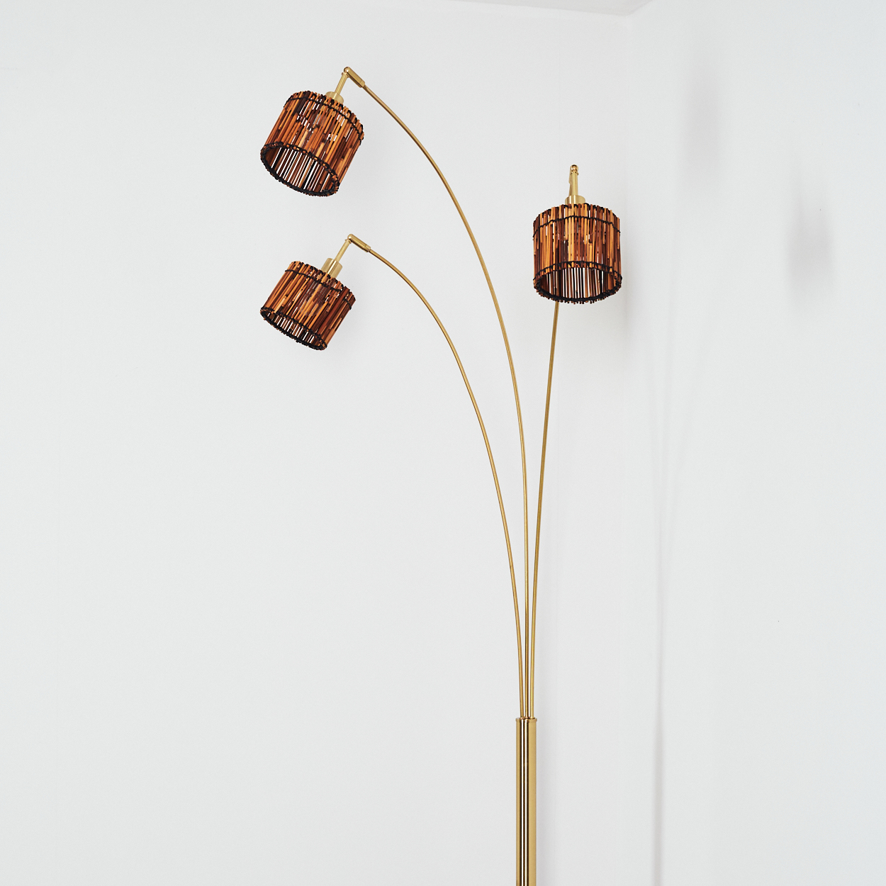 Eine goldene Stehlampe mit drei Lampenschirmen. Die Schirme sind braun und zylindrisch. Der Hintergrund ist eine weiße Wand.
