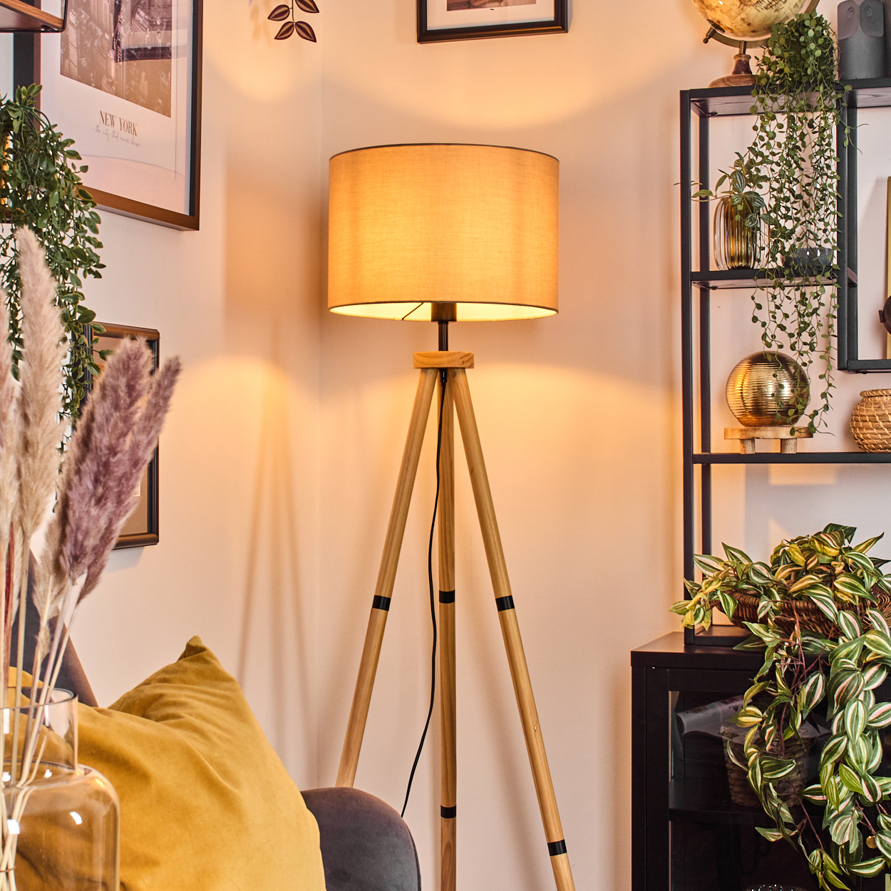 Eine beleuchtete Stativlampe steht in einem Raum. Pflanzen, ein Globus und gerahmte Bilder sind sichtbar. Ein gelbes Kissen und eine Glasvase sind davor.