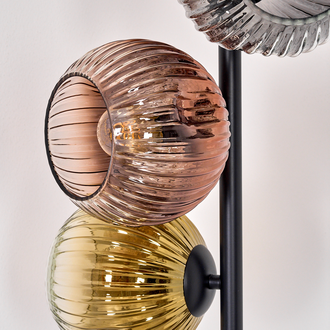 Nahaufnahme einer Lampe mit drei Glasschirmen in Kupfer, Gelb und Klar. Schwarze Stange und weißer Hintergrund.