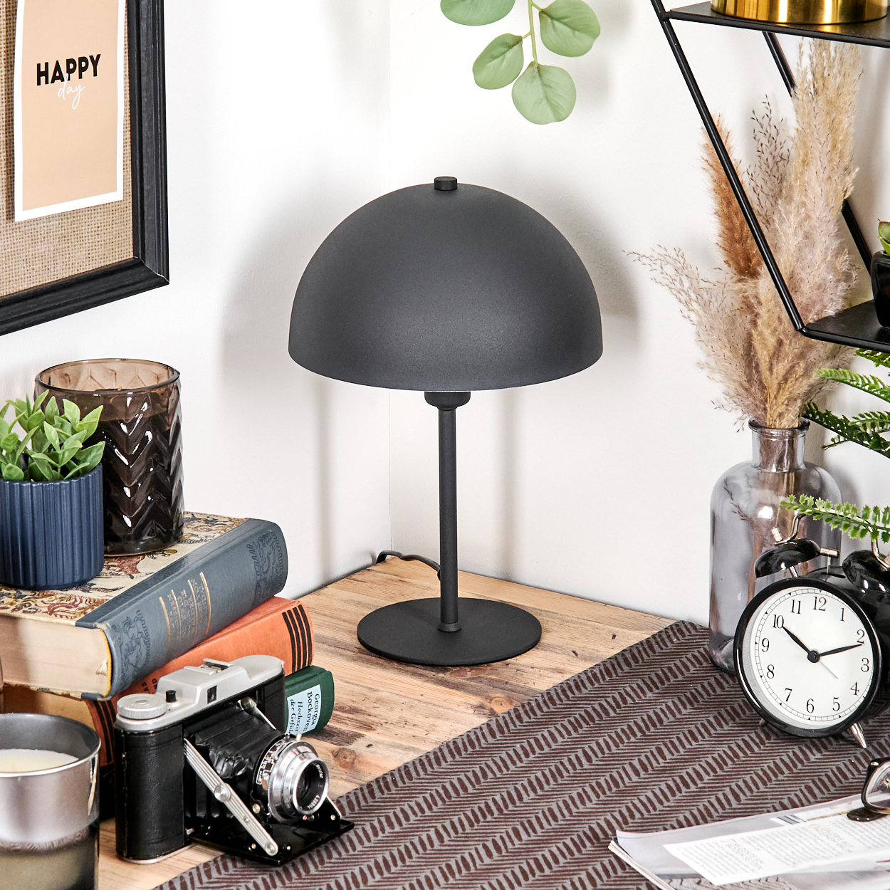 Ein Schreibtisch mit schwarzer Lampe, Büchern, Kamera, Wecker und Pflanzen. Ein gerahmter 'Happy Day'-Druck hängt an der Wand.