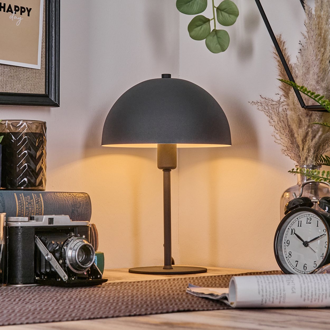 Eine dunkelgraue Lampe steht auf einer Holzoberfläche, neben einer Kamera, Büchern und einem Wecker. Ein aufgerolltes Papier liegt davor.