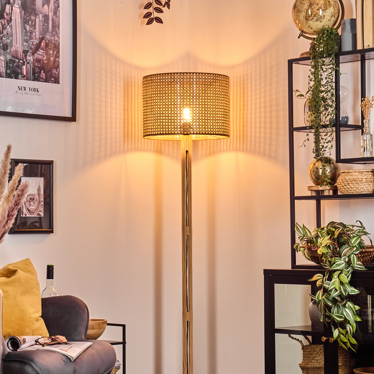 Eine beleuchtete Rattanlampe steht in einem Raum mit einem New York-Poster, einem Samtstuhl und einem schwarzen Regal mit Pflanzen.