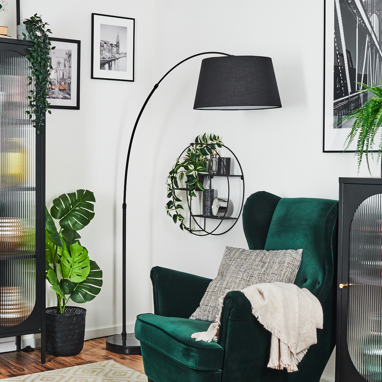 Ein grüner Sessel mit Decke, eine Stehlampe und eine Pflanze stehen vor einer weißen Wand mit Bildern.