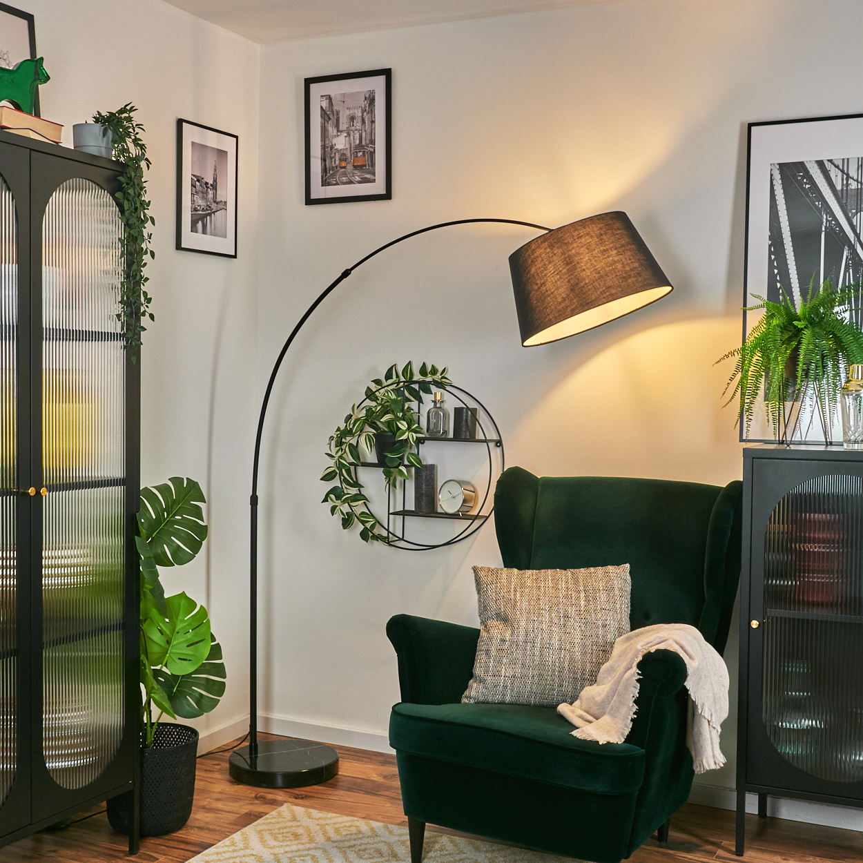 Gemütliches Wohnzimmer mit grünem Sessel, schwarzer Lampe und dekorativen Pflanzen. Bilder an der Wand und ein gemusterter Teppich.
