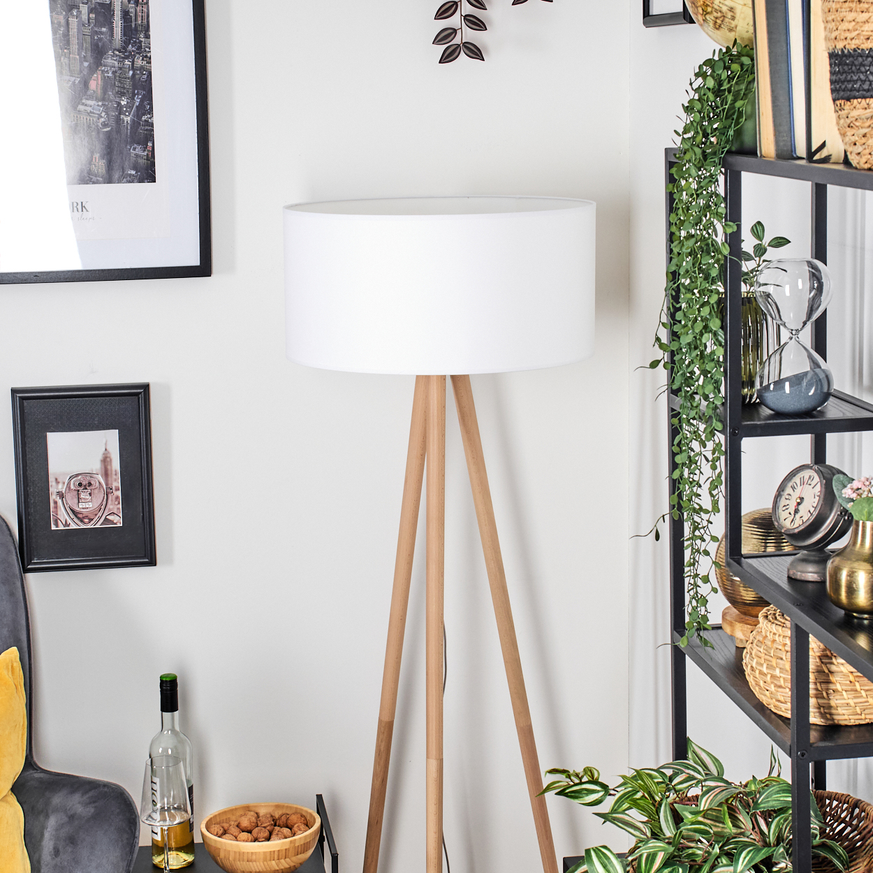Eine weiße Lampe mit Holzbeinen steht vor einem Bücherregal mit Dekoration. Ein Bilderrahmen und eine Flasche sind zu sehen.