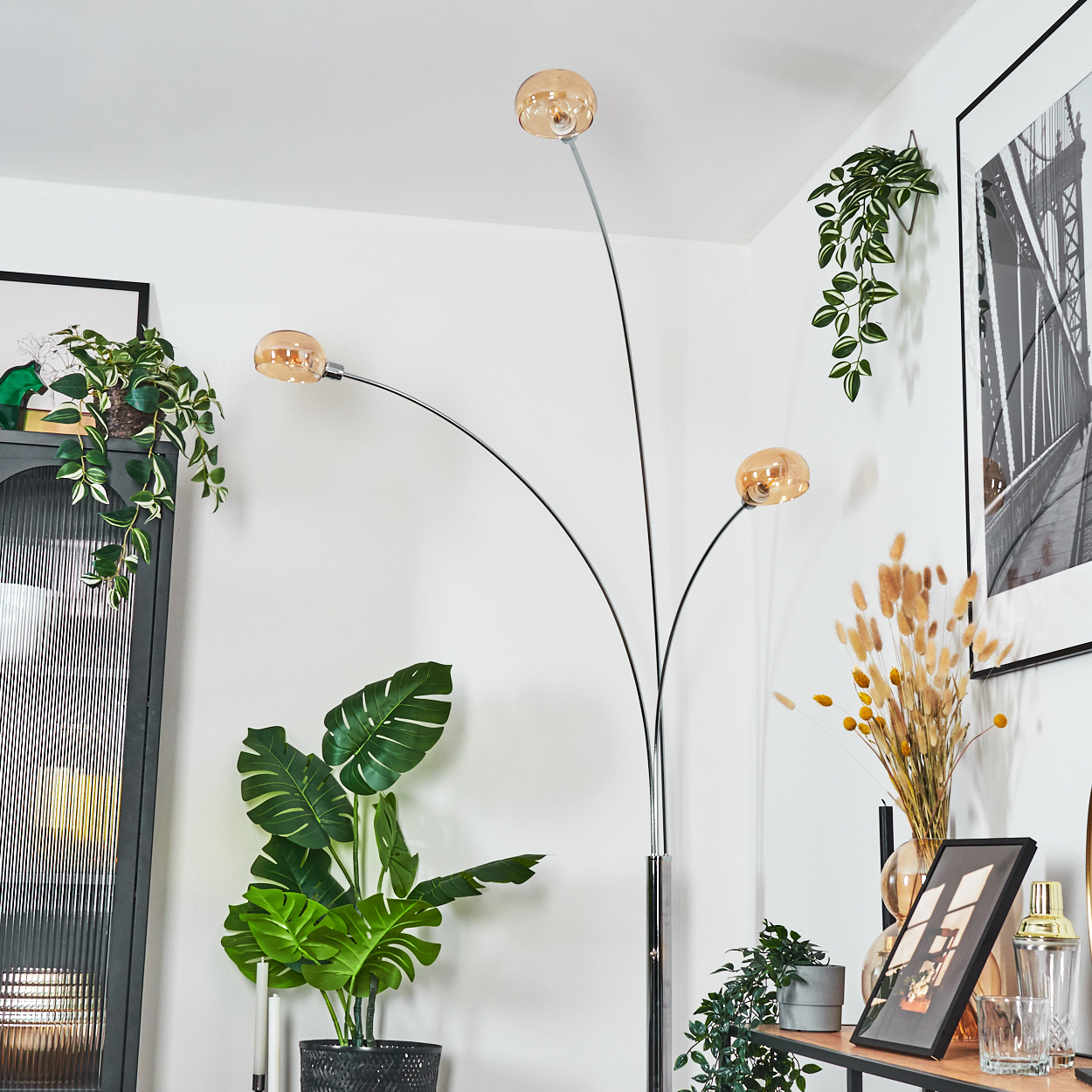 Ein modernes Wohnzimmer mit Stehlampe, Pflanzen und gerahmter Kunst. Die Lampe hat drei bernsteinfarbene Glasschirme. Pflanzen an Wänden.