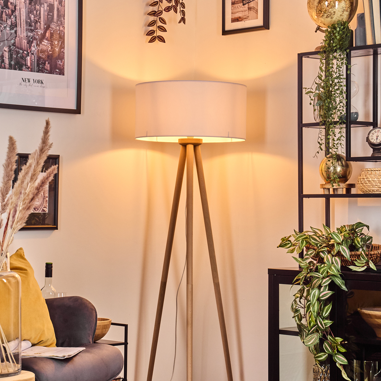 Ein Raum mit einer hölzernen Stativlampe, New York-Poster, Pflanzen und einem schwarzen Metallregal. Im Vordergrund befindet sich eine Couch mit gelbem Kissen.