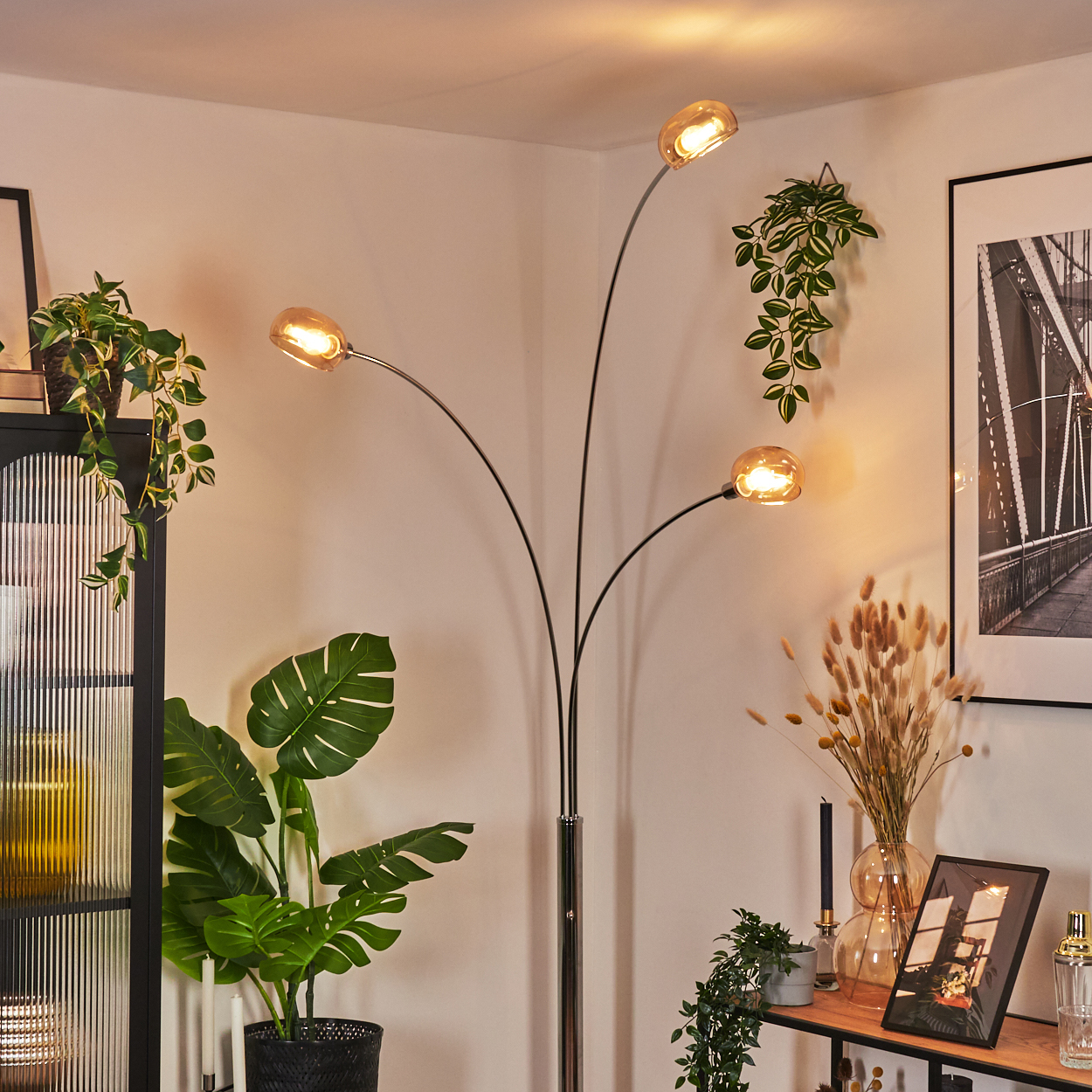 Ein modernes Wohnzimmer mit schwarzer Stehlampe, Pflanzen und einem gerahmten Bild. Die Lampe hat drei Lichter.