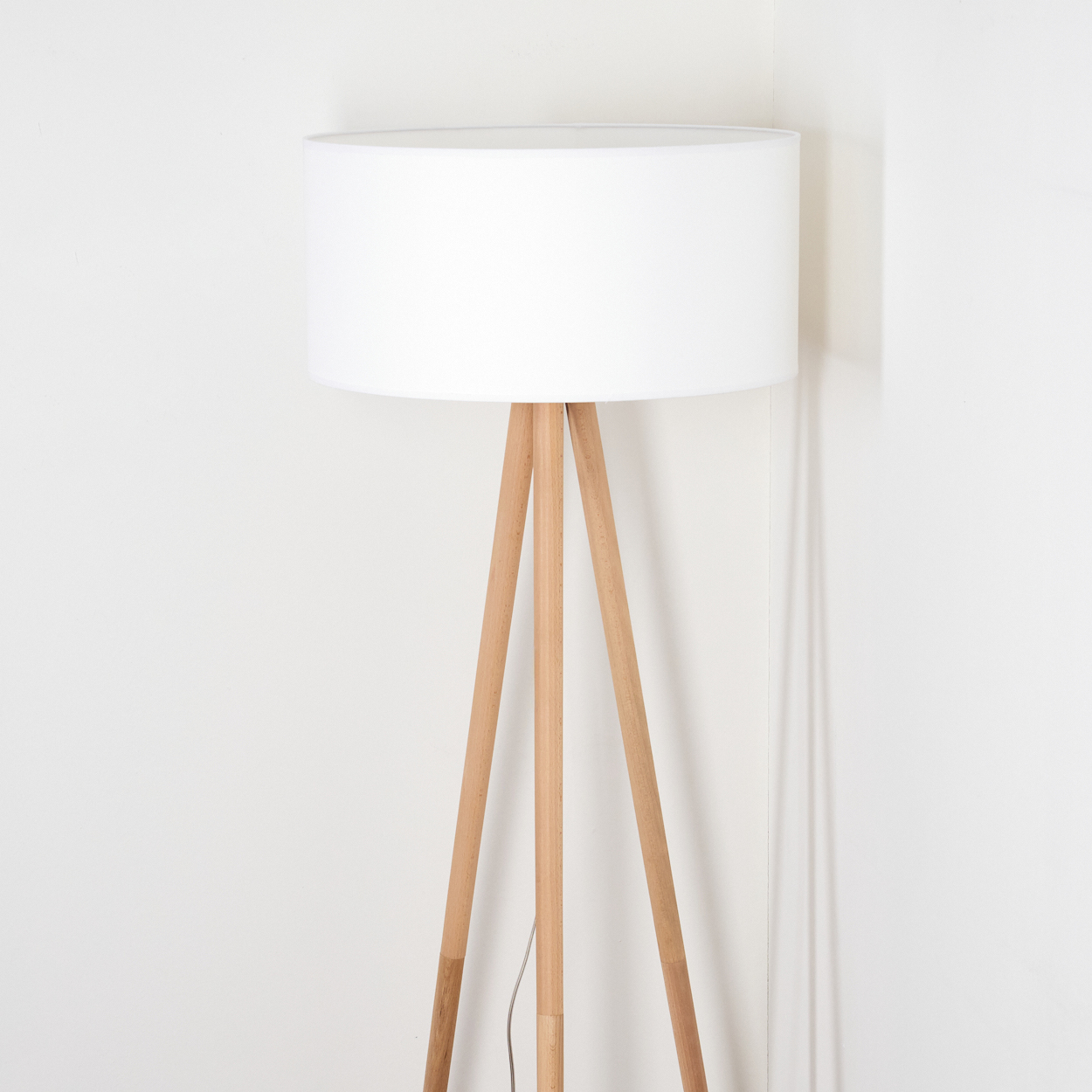 Ein weißer Lampenschirm steht auf Holzbeinen. Die Lampe steht vor einer weißen Wand. Ein Kabel hängt herunter.