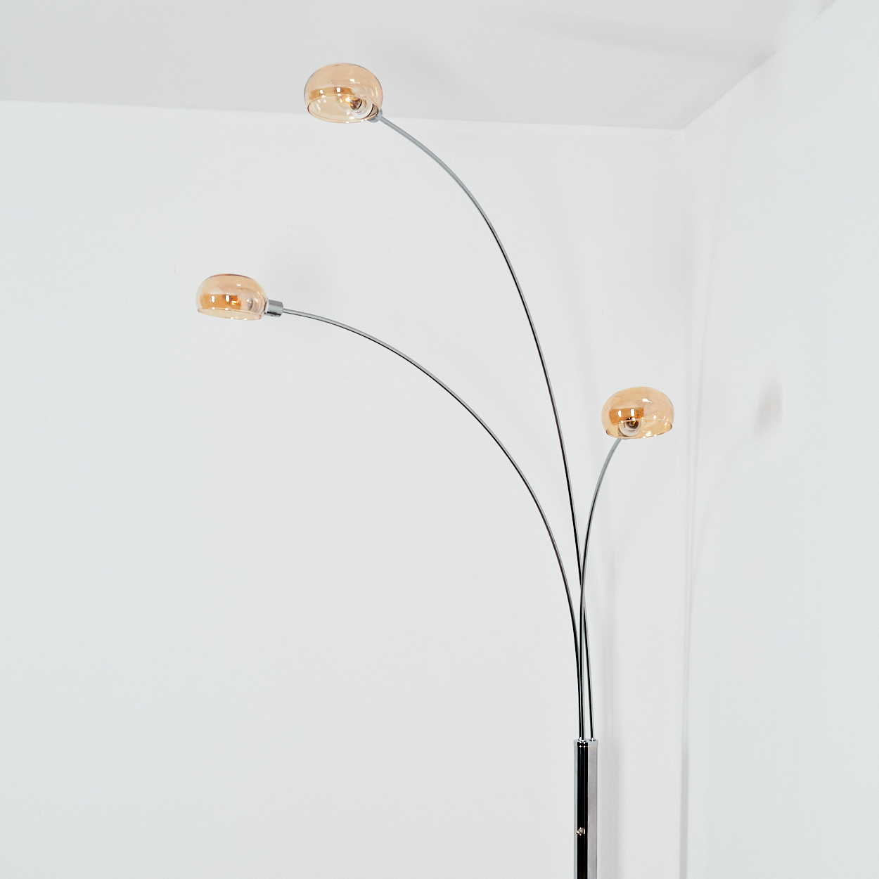 Eine moderne Stehlampe mit drei gebogenen Armen, die jeweils ein rundes, bernsteinfarbenes Licht halten. Sie steht vor einer weißen Wand.