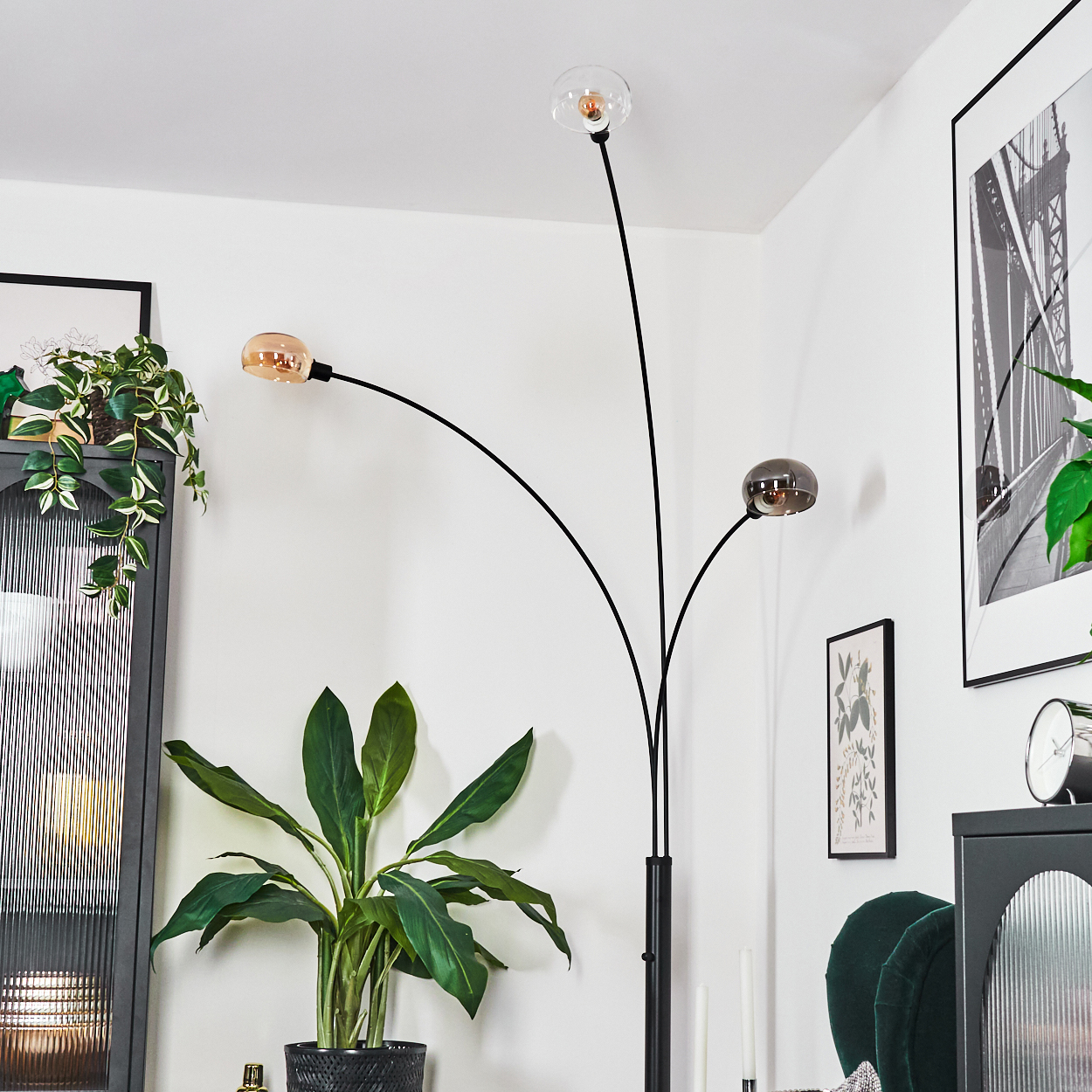 Eine moderne schwarze Stehlampe mit drei Kugellampen. Eine große, belaubte Pflanze befindet sich im Vordergrund, Kunstwerke hängen an den Wänden.