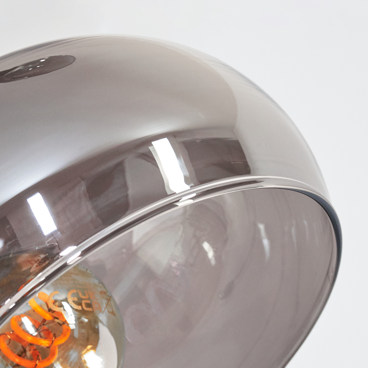 Nahaufnahme einer modernen Lampe mit rauchfarbenem Glasschirm. Im Schirm ist eine spiralförmige, orangefarbene Glühbirne sichtbar. Der Hintergrund ist weiß.