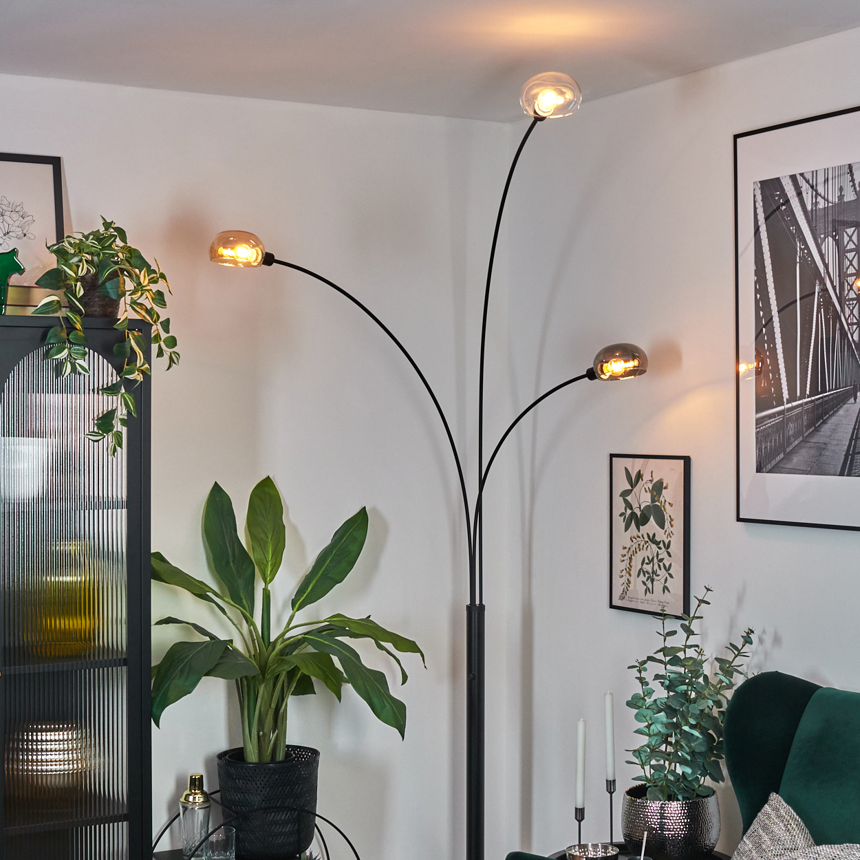 Ein modernes Wohnzimmer mit schwarzer Stehlampe, Pflanzen und gerahmter Kunst. Die Lampe hat drei Lichter. Der Raum hat eine gemütliche Atmosphäre.