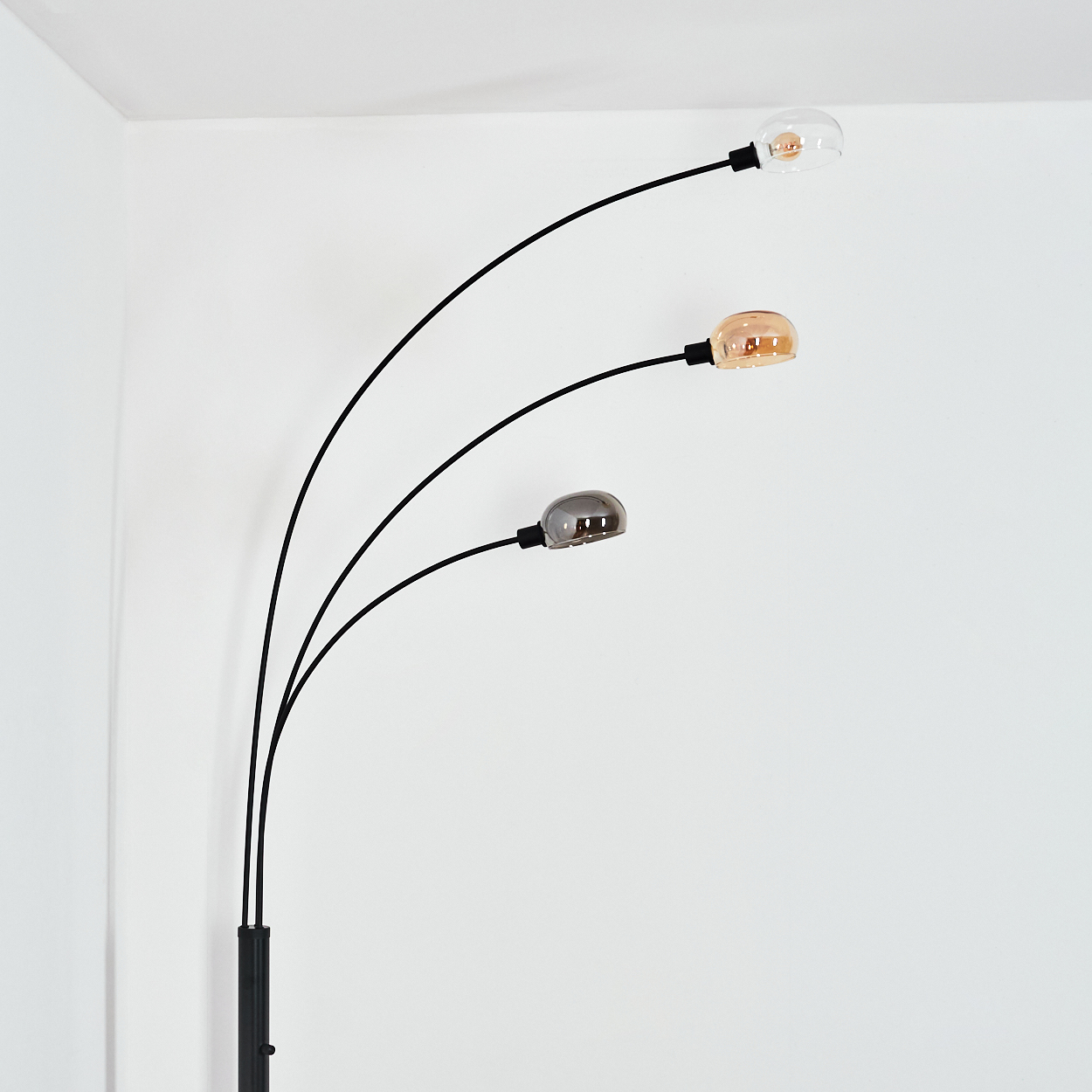 Eine moderne Stehlampe mit drei gebogenen Armen, die jeweils eine Glühbirne mit unterschiedlichen Farbtönen halten: klar, bernsteinfarben und grau. Vor einer weißen Wand.