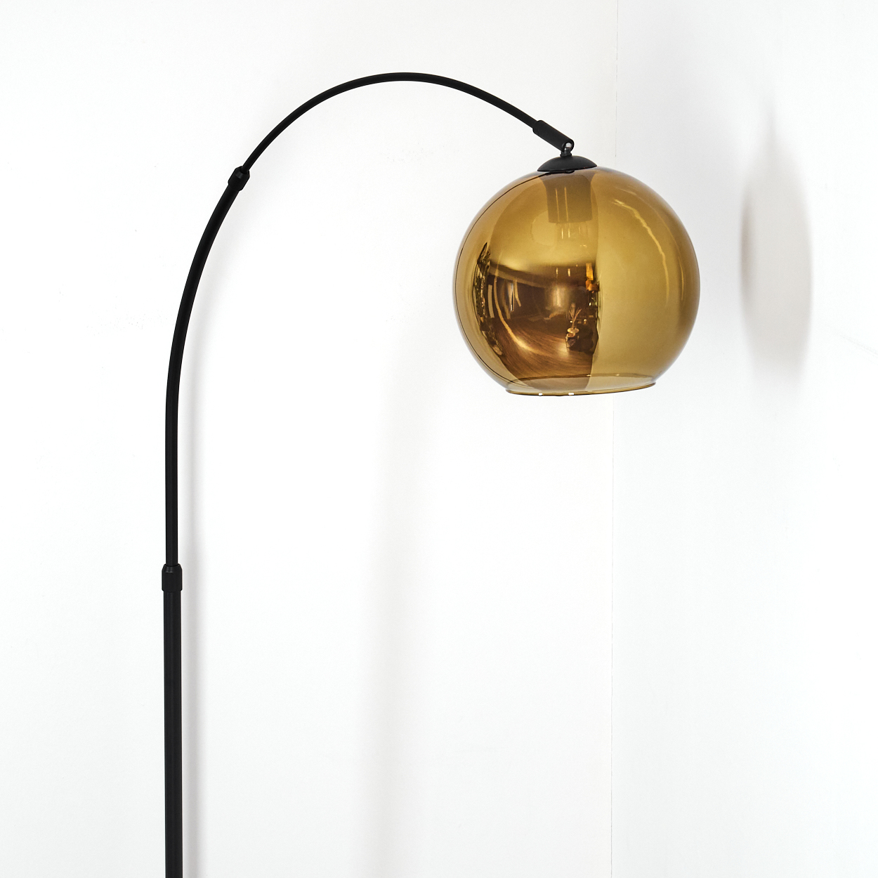 Eine moderne Stehlampe mit schwarzem, gebogenem Arm und goldenem, kugelförmigem Lampenschirm. Der Hintergrund ist eine weiße Wand.