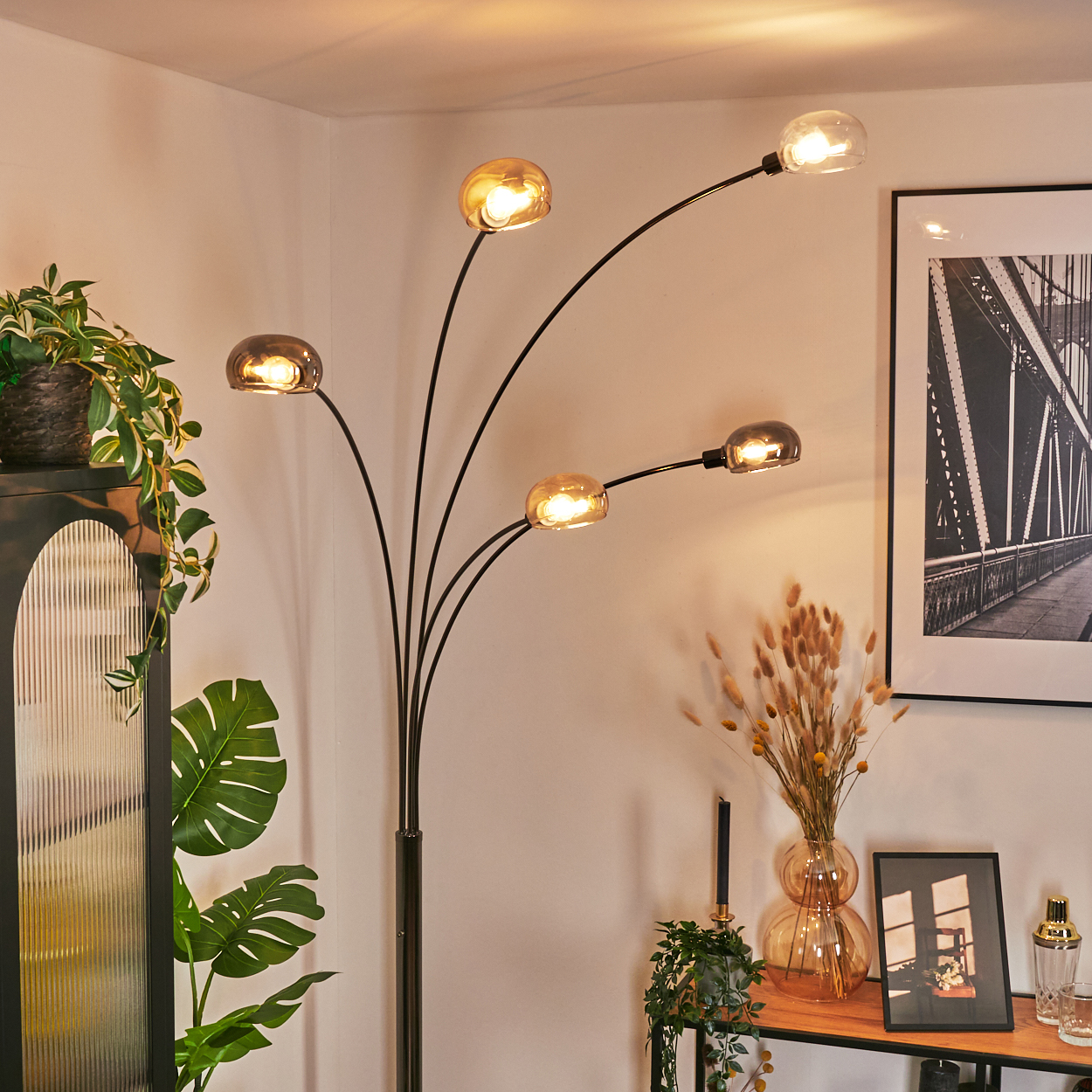 Eine moderne Stehlampe mit fünf Glühbirnen beleuchtet einen Raum. Pflanzen, ein Bild und Dekorationsartikel sind ausgestellt.