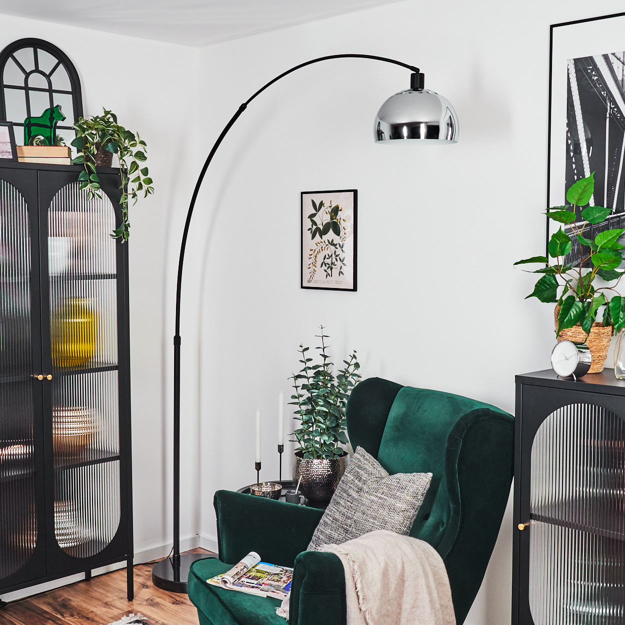 Wohnzimmerszene mit grünem Sessel, schwarzem Schrank, hoher Lampe, Pflanzen und Kunst. Neutrale Töne und eine gemütliche Atmosphäre.