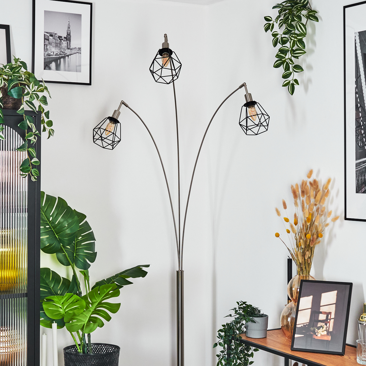 Ein modernes Wohnzimmer mit Stehlampe, Pflanzen, Kunstwerken und einem Regal. Die Lampe hat drei Leuchten mit geometrischen Schirmen.
