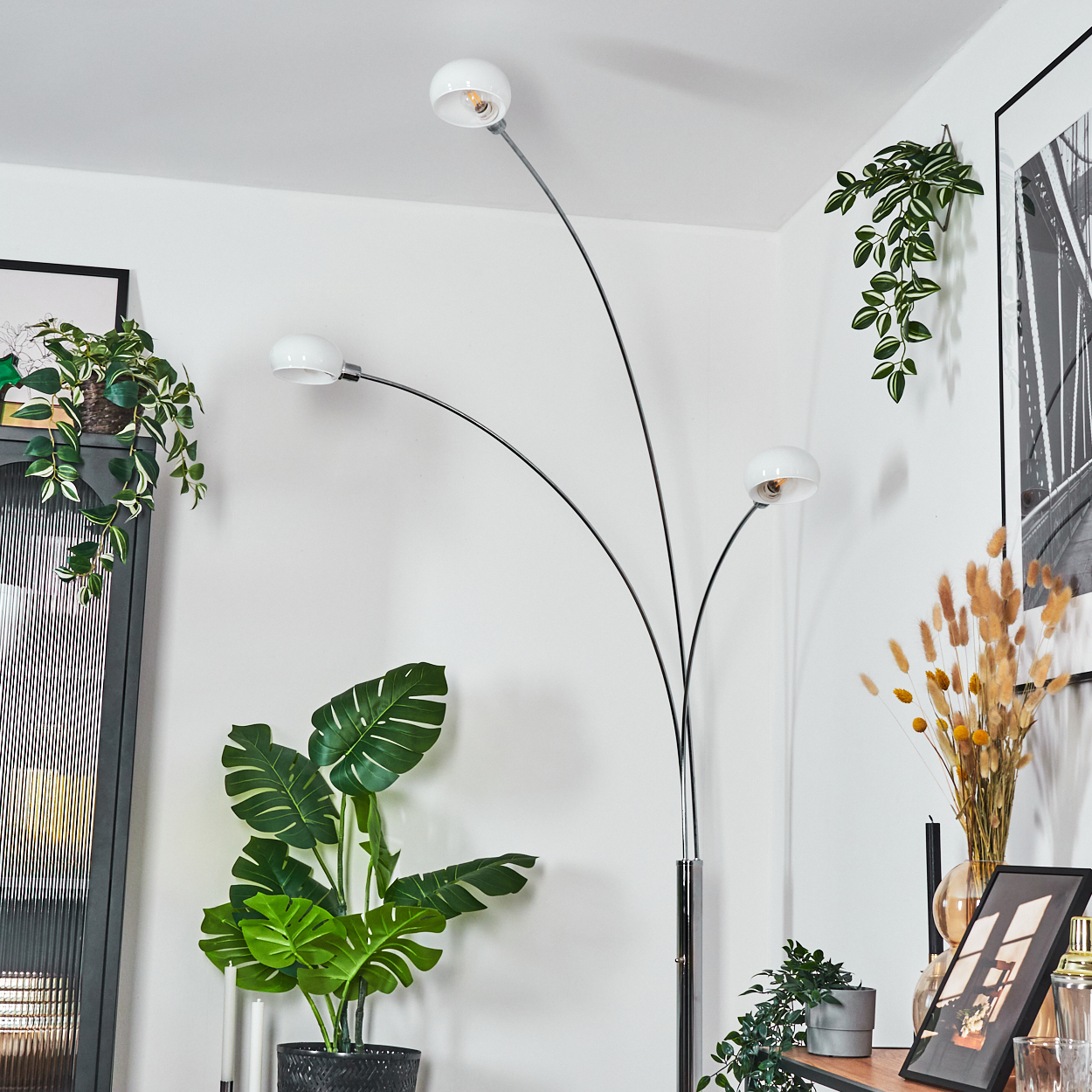 Eine moderne Stehlampe mit drei weißen Lampenschirmen. Topfpflanzen und gerahmte Kunst schmücken eine Ecke eines Raumes.